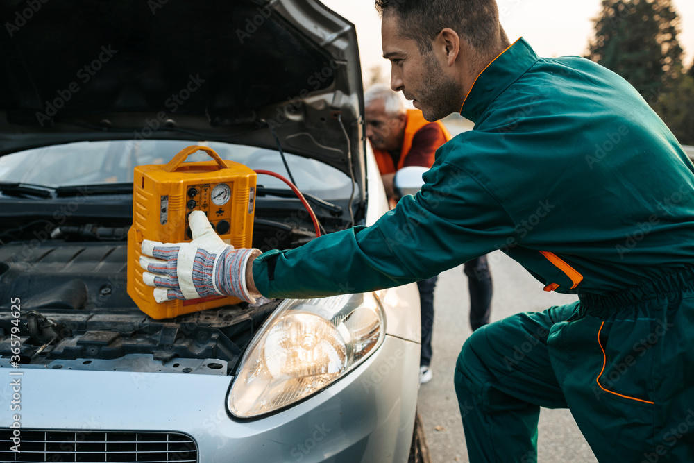Two road assistant workers in towing service trying to start car engine with jump starter and energy station with air compressor. Roadside assistance concept. Two road assistant workers in towing service trying to start car engine with jump starter and energy station with air compressor. Roadside assistance concept.
