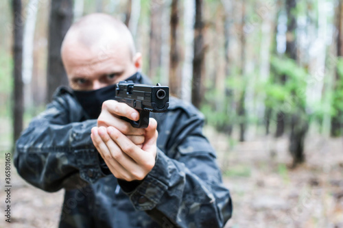A man in dark military clothes and a mask is aiming from a weapon in the forest, close-up