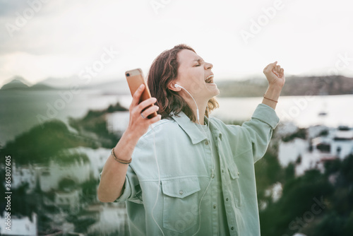 Beautiful happy curly-haired young girl hears music from smartphone in headphones and sings with city view