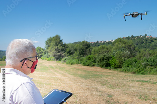 Man with grey hair flying a drone on a field with a tablet and a safety face mask. New normality concept.