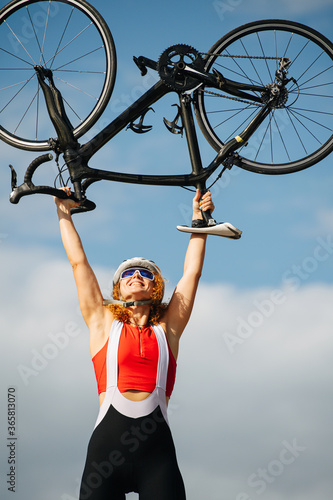 Strong victorious female athlete lifting bike over her head. Low angle