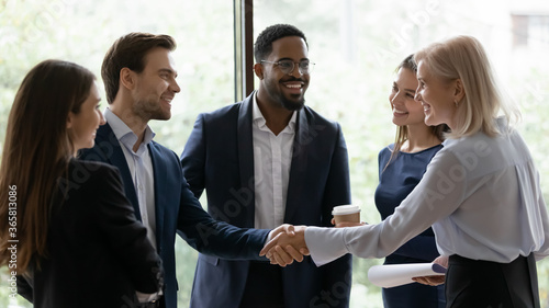 50s business lady shake hands greeting caucasian millennial businessman surrounded by multi ethnic businesspeople in formal suits. Sign contract express trust, partnership, start group meeting concept photo