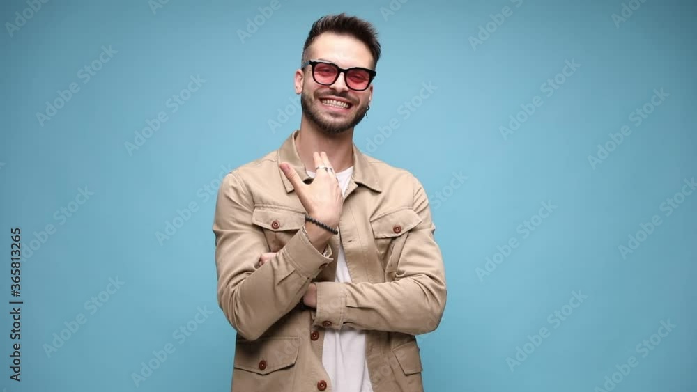 happy young fashion man wearing jacket, smiling and making cutting off sign, moving, holding fists in the air and celebrating victory, looking to side, dancing and shaking fists on blue background happy young fashion man wearing jacket, smiling and making cutting off sign, moving, holding fists in the air and celebrating victory, looking to side, dancing and shaking fists on blue background