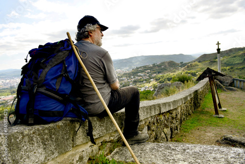 Senior man with backpack and hiking stick looks at a city from a hill photo