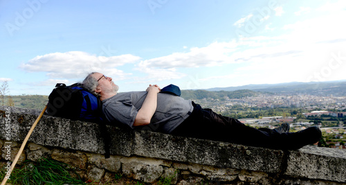 Senior man with backpack and hiking stick rests after hiking on top of a hill photo
