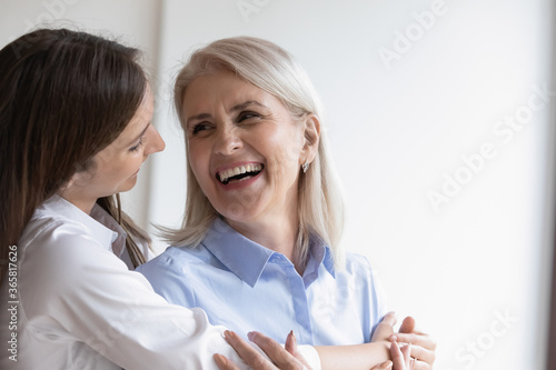 Loving adult daughter hugs cheerful elderly mother close up. Picture of happy grown up child cuddles mommy from behind, pretty women enjoy tender moment looking at each other. Love and warmth concept
