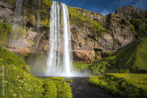 Waterfall Seljalandsfoss falling from high cliff in Iceland and tourists near it. The waterfall drops 60m and is part of the Seljalands River. Visitors can walk behind the falls into a small cave.