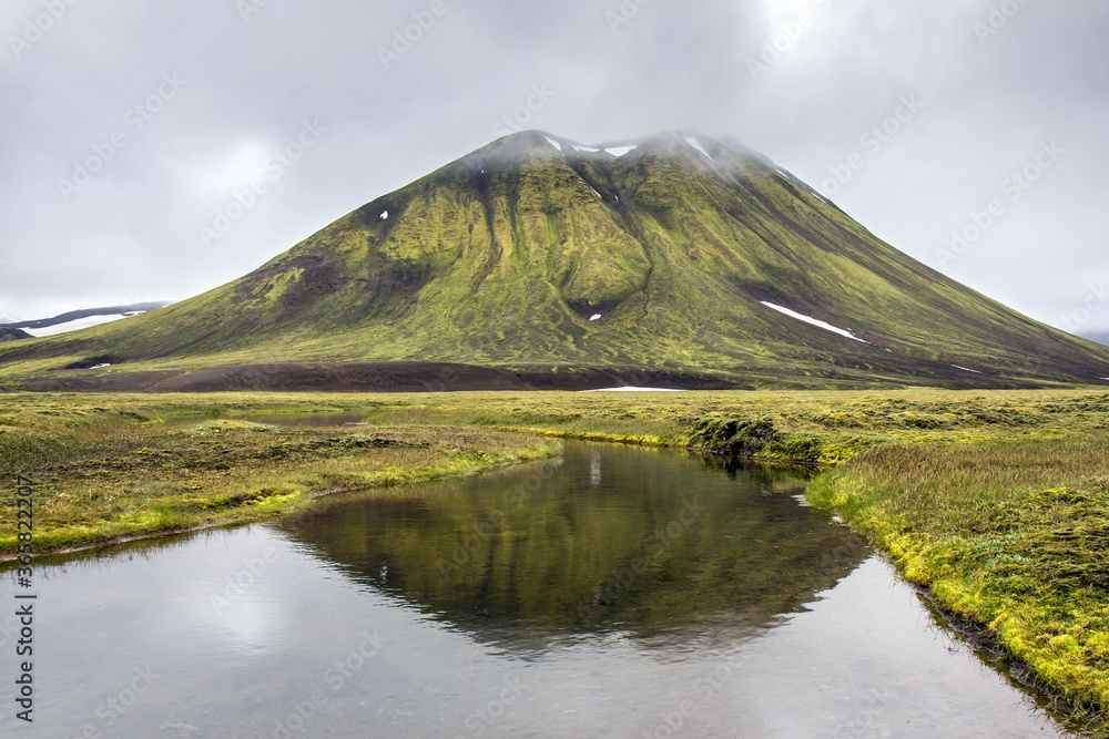 Ancient icelandic volcano and lake on the background. Picturesque reflection of green mountain in lake water. Iceland.