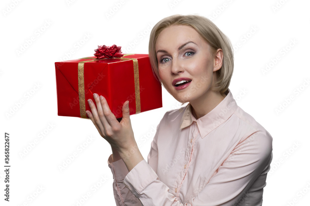 Curious young lady looking up and putting her ear to red gift box. Concept of a expectation of satisfaction from a gift. Curious young lady looking up and putting her ear to red gift box. Concept of a expectation of satisfaction from a gift.