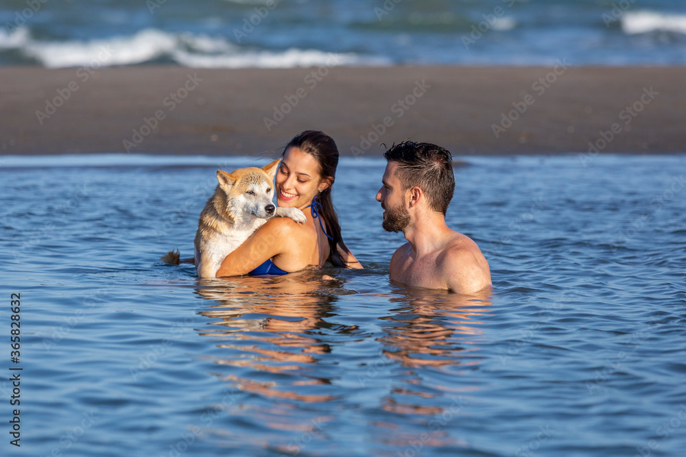 A young pretty couple cuddling and playing with a dog inside the water of a beach A young pretty couple cuddling and playing with a dog inside the water of a beach