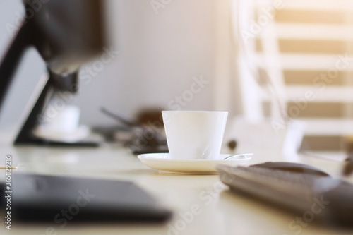 White ceramic cup of hot coffee or tea on wooden computer desk with natural warm sunlight in the morning at office. Caffeine drinks cause a fresh and aromatic feeling.