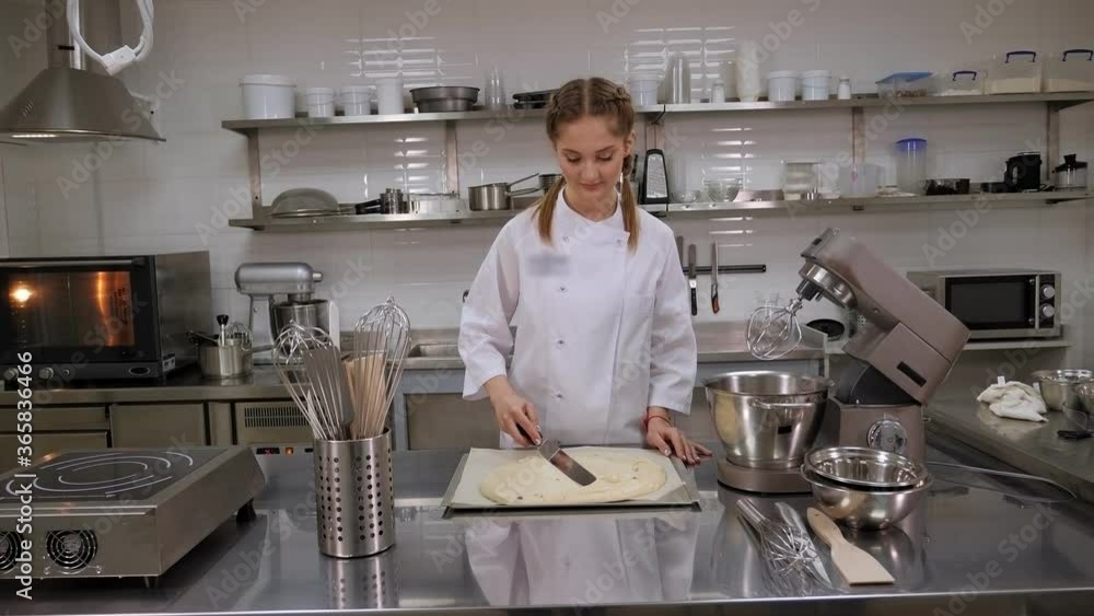 A pastry chef works in a bakery. A female chef in uniform puts a spatula of sponge dough in a baking dish, on the table is a professional food processor. A pastry chef works in a bakery. A female chef in uniform puts a spatula of sponge dough in a baking dish, on the table is a professional food processor.