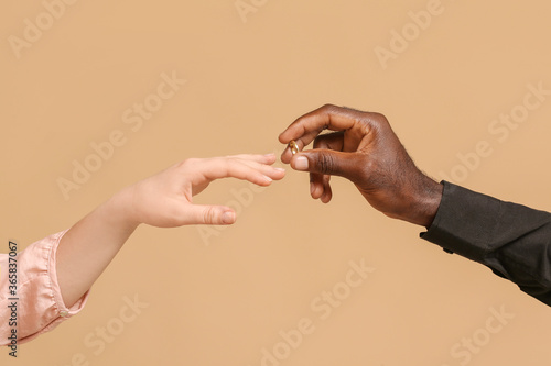 African-American man putting wedding ring on finger of Caucasian woman against color background. Racism concept