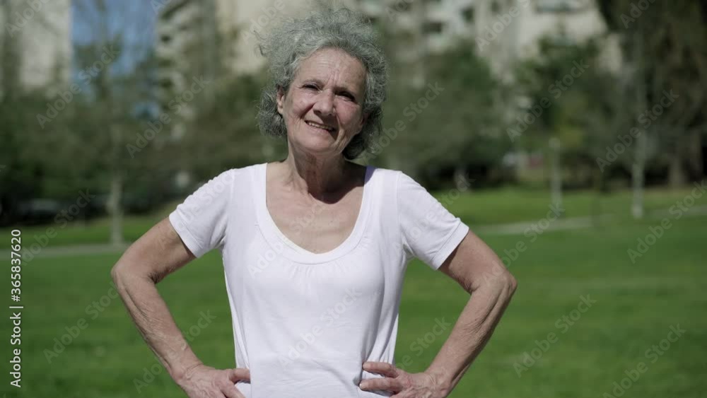 Happy senior woman in sporty t-shirt posing in city park, standing on grass and smiling at camera. Front view. Outdoor fitness activity concept Happy senior woman in sporty t-shirt posing in city park, standing on grass and smiling at camera. Front view. Outdoor fitness activity concept