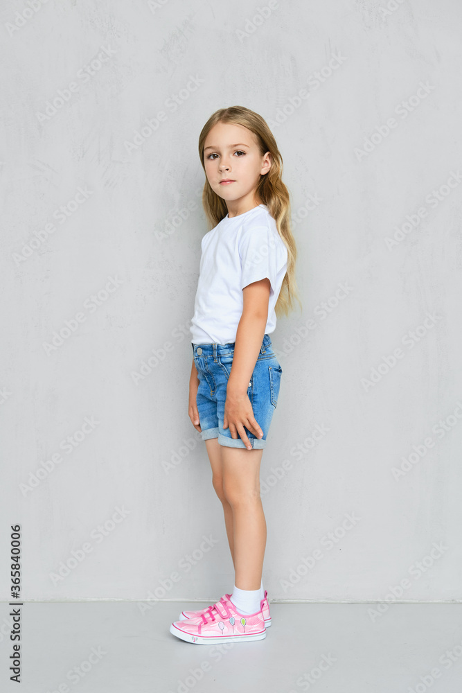 Little child girl in t-shirt, jeans shorts and sneakers posing near the wall Little child girl in t-shirt, jeans shorts and sneakers posing near the wall