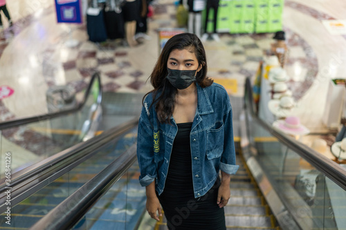 Young Asian woman wearing protective face covering coming up escalator in shopping mall