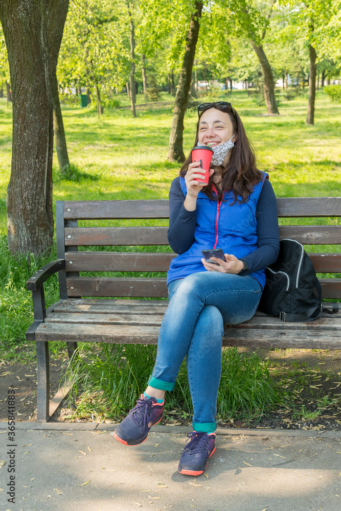 The woman sitting on the bench in the park and wearing a protective mask and using smartphone . Coronavirus concept. toned. vertical photo The woman sitting on the bench in the park and wearing a protective mask and using smartphone . Coronavirus concept. toned. vertical photo