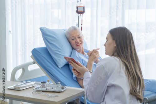 Happy woman doctor talking and using tablet to sick old female senior elderly patient lying in bed in hospital ward room in medical, technology and healthcare treatment concept. Caucasian people. photo