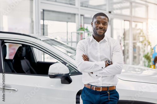 portrait of smiling afro man in white shirt, he stands next to new car, look at camera.