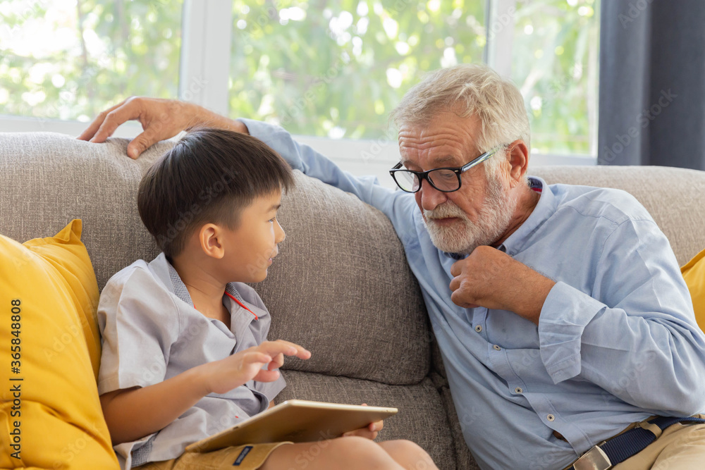 Obraz premium Grandfather and little cute grandson playing touchscreen tablet PC together on sofa in their house