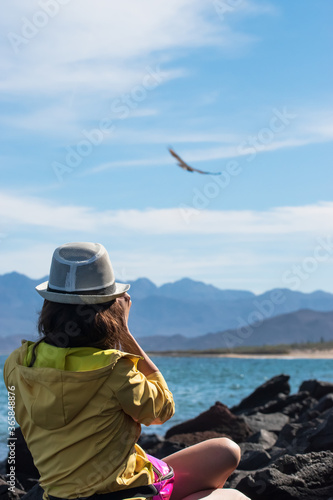 Photographer girl taking pictures of a bird in flight with wide outstretched wings in the sky on the landscape of mountain range in Baja California in Mexico.