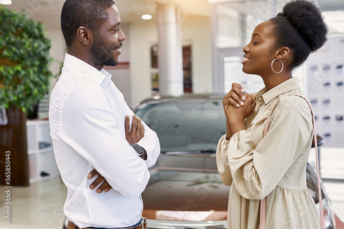 pretty african woman asks her husband to buy a car, she looks at him with love, man smile and think. isolated in car dealership
