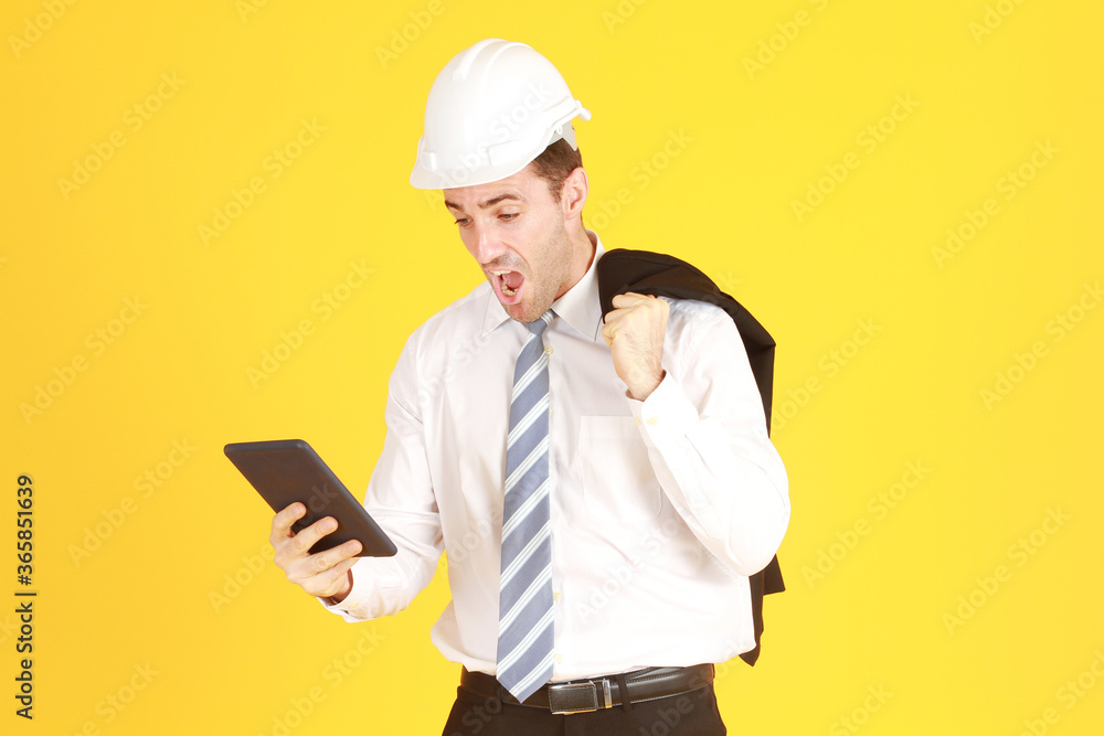 Handsome and smart engineer in suit and white shirt and Wearing a white safety engineering hat with hand holding smartphone Surprised and stressed with work isolated on yellow background. Handsome and smart engineer in suit and white shirt and Wearing a white safety engineering hat with hand holding smartphone Surprised and stressed with work isolated on yellow background.