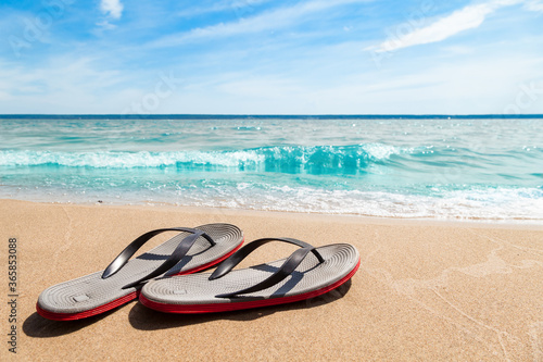 A pair of sandals with red soles on a sunny beach with sand near the seashore on a summer day. Rest and travel to hot countries.