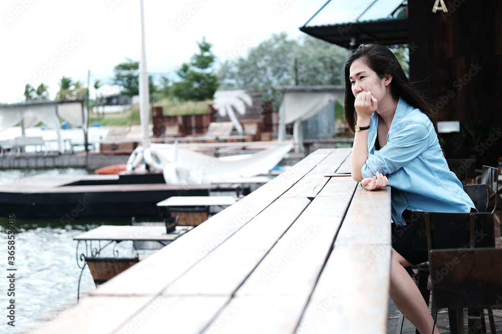 Asian woman long hair and wear blue jean jacket sitting alone on stool chair and wooden bar on terrace near the lake and river.