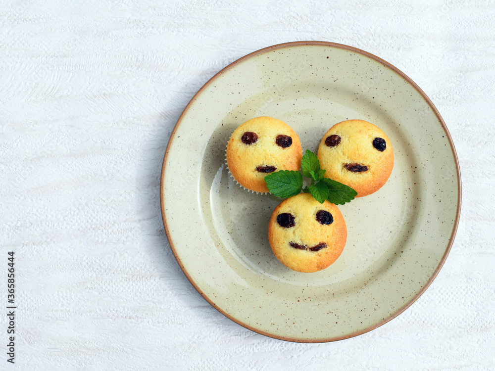 Freshly baked homemade muffins with funny faces. Vanilla muffins with raisins, decorated with melissa leaves Freshly baked homemade muffins with funny faces. Vanilla muffins with raisins, decorated with melissa leaves