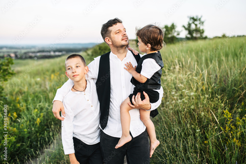 Proud father with two sons walk outdoors. Dad with toddler in his arms and preteen son stand in the scenic field Proud father with two sons walk outdoors. Dad with toddler in his arms and preteen son stand in the scenic field
