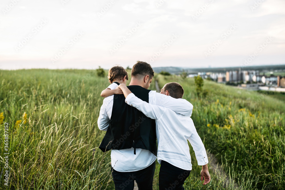 Proud father with two sons walk outdoors. Back view dad with toddler in his arms hugs preteen son, they walk together in a scenic field