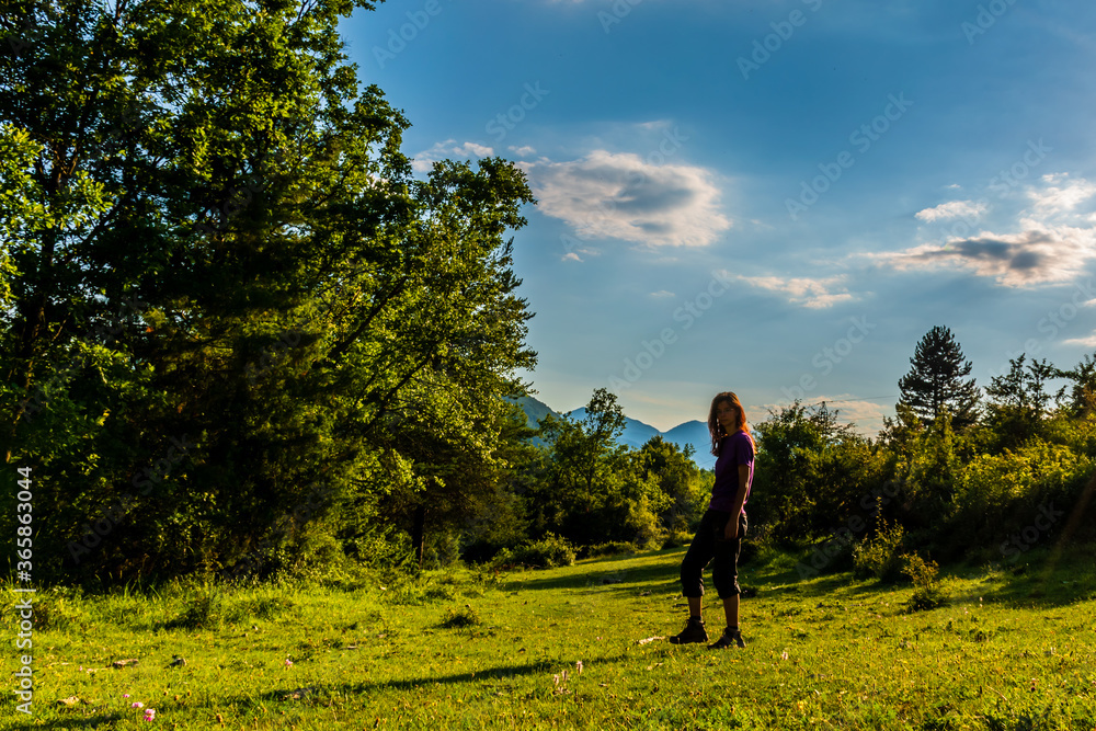 A full body shot of a young Caucasian redhead hiker standing in a meadow in a forest in the French Alps during sunset