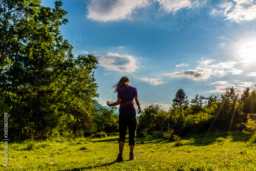 A full body shot of an unrecognizable young Caucasian redhead woman standing in a meadow in a forest in the French Alps during sunset
