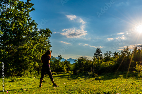 A full body shot of an unrecognizable young Caucasian redhead woman in headphones dancing in a meadow in a forest in the French Alps during sunset