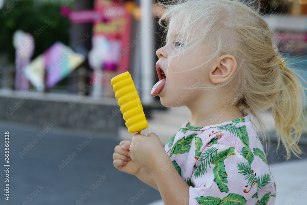 Cute toddler girl eating yellow popsicle. Candid outdoor portrait of child with ice cream. Cute toddler girl eating yellow popsicle. Candid outdoor portrait of child with ice cream.