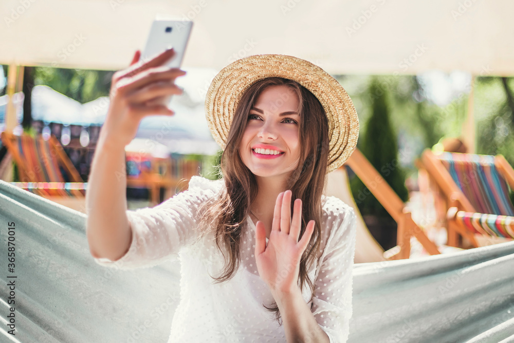 Young woman tourist traveler smiling, waving hand at the phone screen, sitting in a hammock on the beach and making video calls, video conference during the coronavirus quarantine