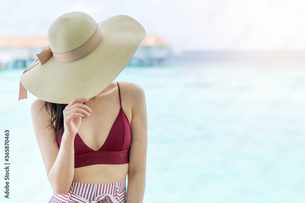 Young girl holding hat walking surrounded by beautiful turquoise ocean water. Happy traveller in tropical beach vacation. Back of young Asian woman walking over a wooden jetty in morning sunrise.