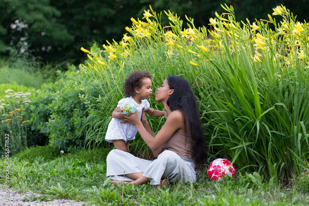 Fototapeta premium Full length horizontal portrait of cute toddler girl bending to kiss her pretty mixed-raced young mother sitting in exterior garden location