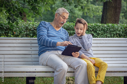 portrait of grandfather and grandson in the park in summer with a tablet in his hands, an elderly man plays games online, the grandson teaches his grandfather to use the Internet