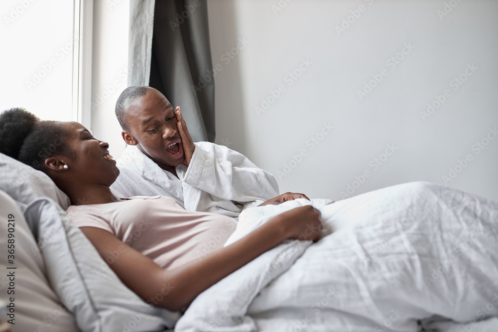 surprised african young man looking at his beutiful pregnant wife lying on bed. he is shocked by the size of his stomach surprised african young man looking at his beutiful pregnant wife lying on bed. he is shocked by the size of his stomach