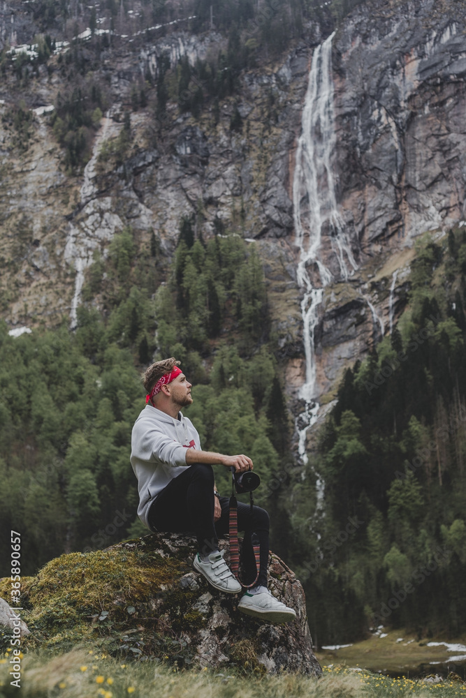 Obraz premium Obersee. Schonau am Konigssee. Bavaria. Germany. Young hipster photographer with camera sits on the stone & looks at Rothbach waterfall on background of pine forest & rock mountain cliff