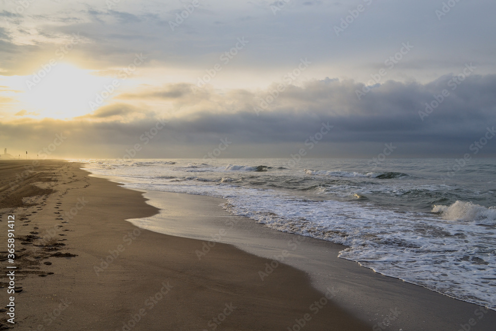Gava Playa beach in Castelldefels, Barcelona, Spain. Bright sun, dark clouds, beautiful coastline. Large waves and fog in the background. Summer morning in nature, sunrise.