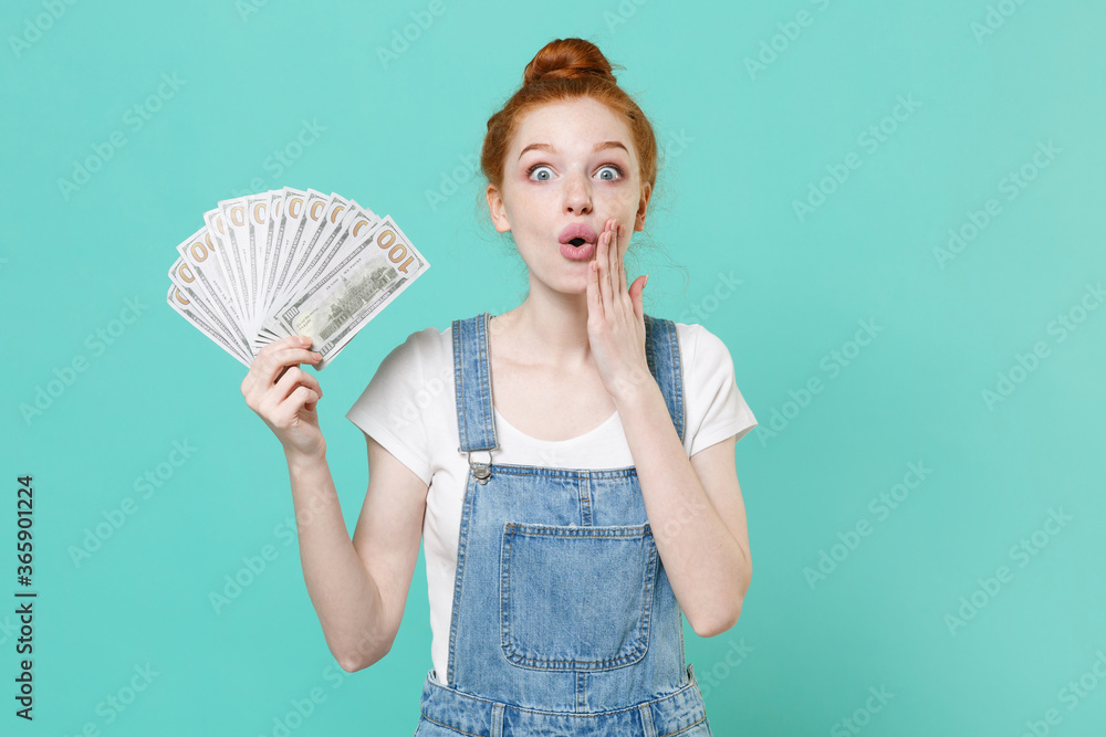 Amazed young readhead girl in casual denim clothes isolated on blue turquoise background. People lifestyle concept. Mock up copy space. Hold fan of cash money in dollar banknotes, put hand on cheek. Amazed young readhead girl in casual denim clothes isolated on blue turquoise background. People lifestyle concept. Mock up copy space. Hold fan of cash money in dollar banknotes, put hand on cheek.