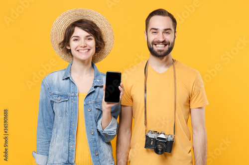 Smiling tourists couple friends guy girl with photo camera isolated on yellow background. Passenger traveling abroad on weekends. Air flight journey concept. Hold mobile phone with blank empty screen.