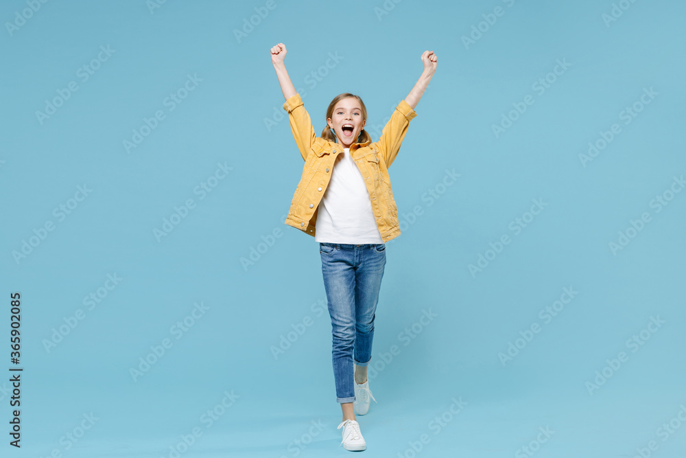Full length portrait of excited little blonde kid girl 12-13 years old in yellow jacket isolated on blue wall background. Childhood lifestyle concept. Mock up copy space. Clenching fists like winner.