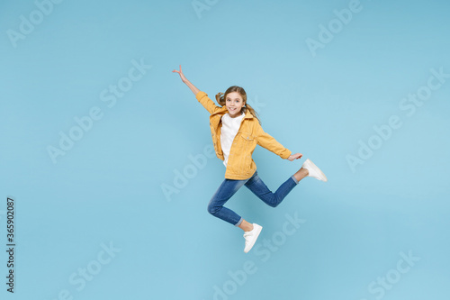 Full length portrait of smiling little blonde kid girl 12-13 years old in yellow jacket isolated on blue background studio. Childhood lifestyle concept. Mock up copy space. Jumping, spreading hands.