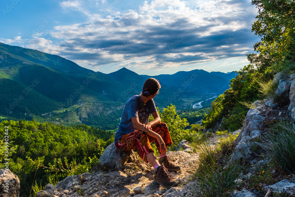 A full body shot of a thoughtful unrecognizable young Caucasian redhead hiker sitting on a boulder on a hiking path and enjoying the view of the French Alps during sunset