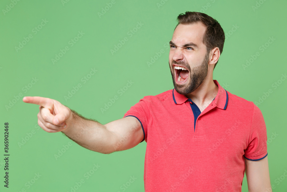 Irritated young bearded man guy in casual red pink t-shirt posing isolated on green background studio. People lifestyle concept. Mock up copy space. Pointing index finger aside screaming swearing. Irritated young bearded man guy in casual red pink t-shirt posing isolated on green background studio. People lifestyle concept. Mock up copy space. Pointing index finger aside screaming swearing.