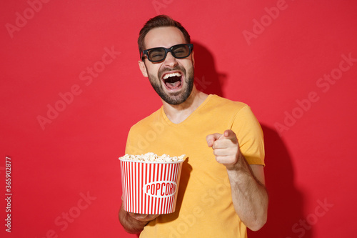 Laughing young man guy in casual yellow t-shirt 3d glasses isolated on red background. People in cinema, lifestyle concept. Watching movie film holding bucket of popcorn point index finger on camera.